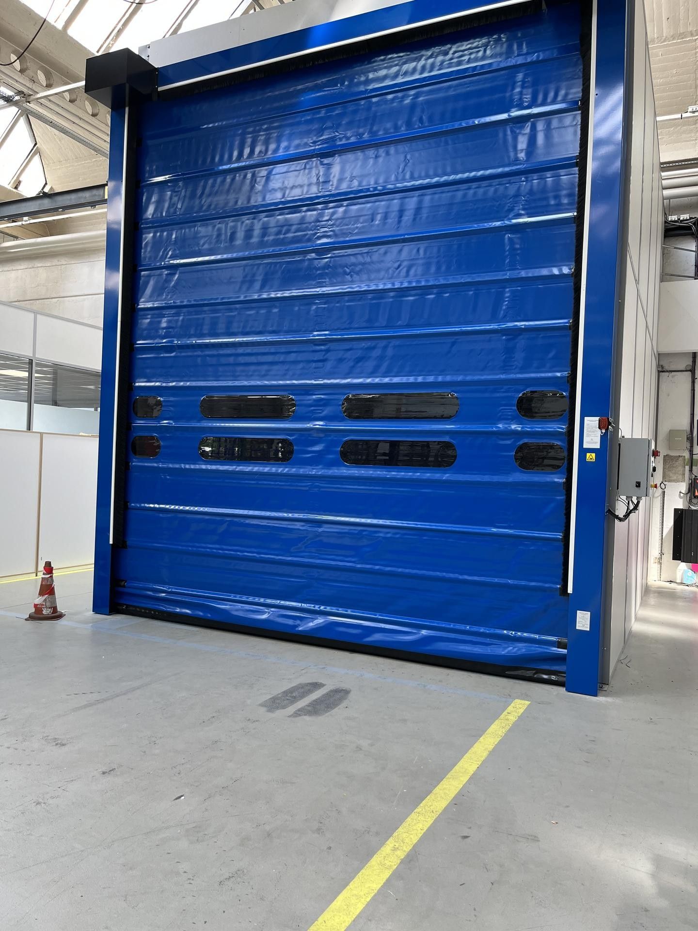 Porte rapide souple bleue à hublots dans un hall industriel.